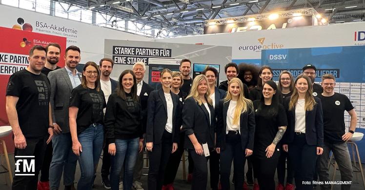 Eine große Gruppe junger Menschen steht vor einem Messestand. Auf den Wänden stehen Texte wie 'Starker Partner', 'DSSV' und 'Spitzen Firmen der Branche'. Alle tragen sportlich-elegante Kleidung.