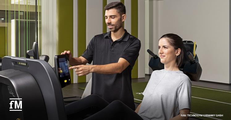 Ein Trainer erklärt einem Mitglied die Nutzung eines Technogym-Geräts für personalisierte Trainingspläne