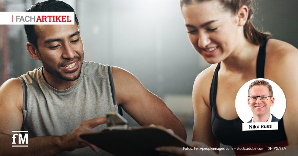 Ein Trainer mit zeigt einer jungen Frau ein Klemmbrett. Beide lächeln und sitzen in einem Fitnessstudio.