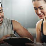 Ein Trainer mit zeigt einer jungen Frau ein Klemmbrett. Beide lächeln und sitzen in einem Fitnessstudio.