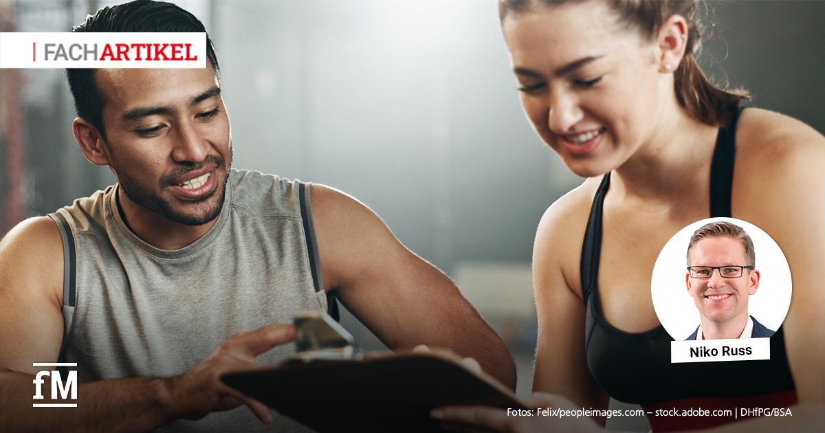 Ein Trainer mit zeigt einer jungen Frau ein Klemmbrett. Beide lächeln und sitzen in einem Fitnessstudio.
