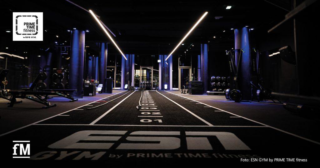 Modern ausgestatteter Functional-Training-Bereich mit Sprintbahn und weißem Schriftzug 'ESN GYM by PRIME TIME fitness', Fitnessgeräten und dunkler, lilablauer Studiobeleuchtung.