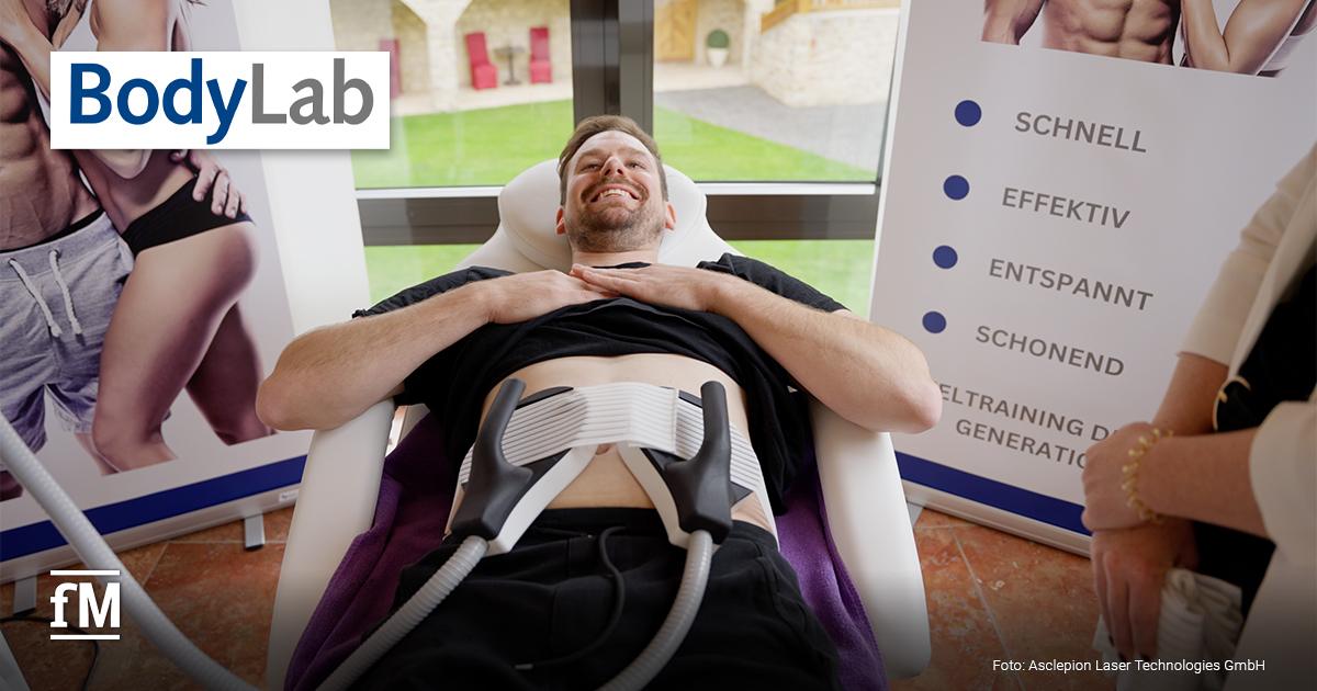 Ein Mann liegt auf einer Liege und lächelt. Zwei Bodyforming-Applikatoren sind an seinem Bauch befestigt. Im Hintergrund sind Poster mit Fitnessmotiven und Informationen zur Behandlung zu sehen.