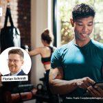 Ein junger Mann im Sportoutfit steht in einem modernen Fitnessstudio und bedient ein Tablet mit einem Lächeln im Gesicht. Im Hintergrund trainiert eine Frau an einem Boxsack vor einer Wand aus roten Backsteinen. Oben links im Bild ist der Hinweis 'FACHARTIKEL' zu sehen. Darunter befinden sich zwei Porträtfotos: links Nicolai Rolli, rechts Florian Schmidt.