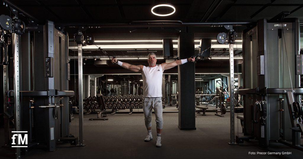 Ein Mann trainiert mit ausgestreckten Armen an einem doppelten Kabelzuggerät in einem modernen Fitnessstudio mit Hanteln im Hintergrund.