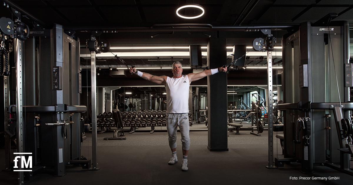 Ein Mann trainiert mit ausgestreckten Armen an einem doppelten Kabelzuggerät in einem modernen Fitnessstudio mit Hanteln im Hintergrund.