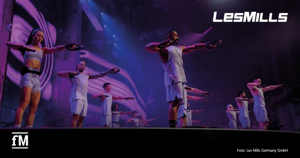 Mehrere Les Mills Trainer präsentieren auf einer großen Bühne eine synchronisierte Fitness-Choreografie mit speziellen Hantelscheiben. Die Teilnehmer tragen helle, sportliche Outfits. Im Hintergrund sorgen dynamische Lichteffekte und LED-Wände für eine beeindruckende Atmosphäre. Das Les Mills Logo ist oben rechts zu sehen, das fM-Logo unten links.