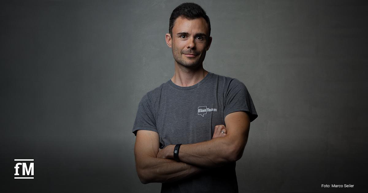Ein Mann steht mit verschränkten Armen vor dunklem Hintergrund. Er trägt ein graues T-Shirt mit dem Aufdruck 'Elan Team' und blickt ruhig und selbstbewusst in die Kamera.