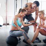 Im Vordergrund erklärt eine Trainerin in schwarzem Poloshirt einer Kundin die Trainingsdaten auf einem großen Touchscreen im Fitnessstudio. Im Hintergrund sind moderne Geräte zur Bewegungsförderung zu sehen. Links im oberen Bilddrittel ist ein rundes Porträt des Autors Andreas Barz zu sehen, darüber steht 'Fachartikel', der erste Teil in grauer, der zweite Teil in roter Schrift.