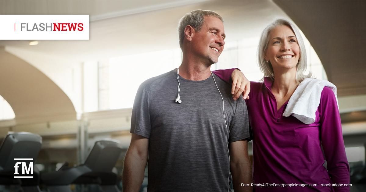 Ein sportliches, älteres Paar steht entspannt in einem modernen Fitnessstudio. Der Mann trägt ein graues Funktionsshirt mit Kopfhörern um den Hals, die Frau ein lila Sportoberteil mit einem Handtuch über der Schulter. Beide lächeln sich glücklich an. Im Hintergrund sind Laufbänder und große Fenster zu sehen. Oben links im Bild steht 'FLASH NEWS' in Rot und Schwarz, unten links befindet sich das fM-Logo.