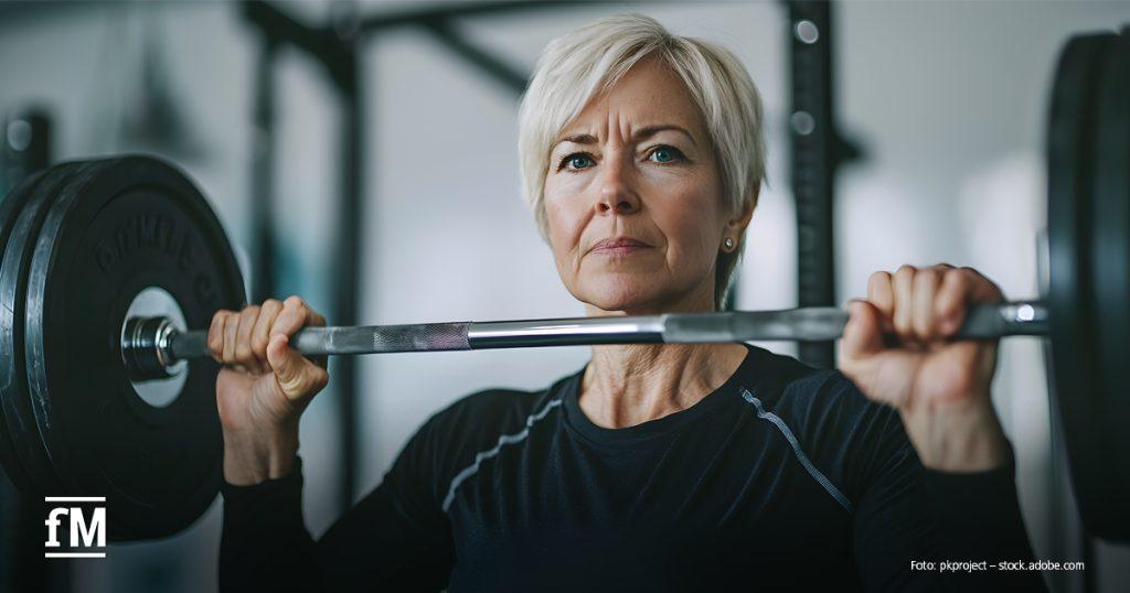 Eine ältere Frau hebt konzentriert eine Langhantel im Fitnessstudio. Sie trägt ein schwarzes Sportshirt. Ihr Blick ist fokussiert.