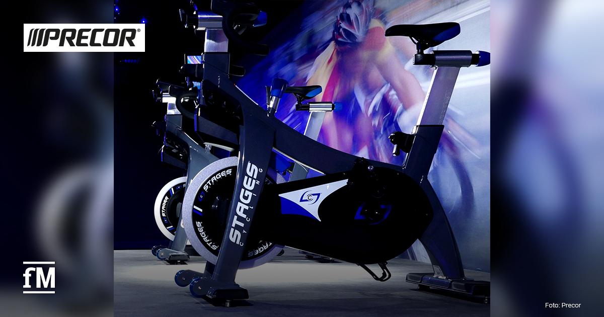 Indoor-Cycling-Bikes von Stages im Ausstellungsraum, beleuchtet mit blauen und weißen Akzenten. Logo von Precor und Stages sind sichtbar.