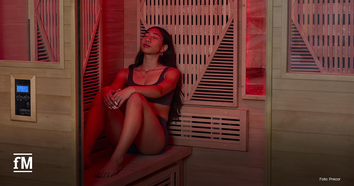 Eine junge Frau sitzt entspannt in einer modernen Sauna mit rotem Licht. Sie trägt einen roten Sport-BH und eine passende Leggings. Das Logo von Precor ist unten links zu sehen.