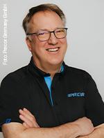 Ein Mann mit Brille und kurzen grauen Haaren lächelt freundlich. Er trägt ein schwarzes Poloshirt mit Precor-Logo und steht vor einem neutralen Hintergrund.