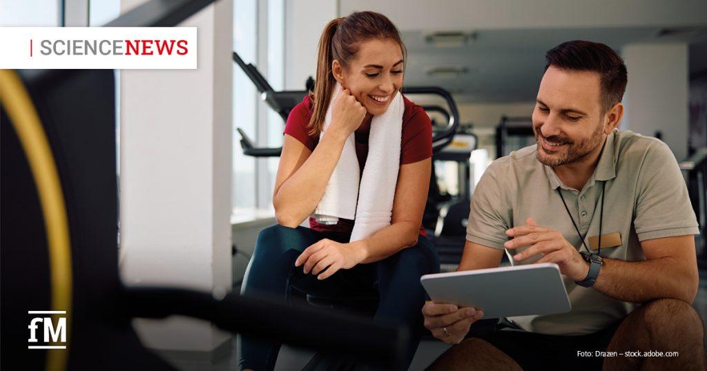 Eine junge Frau mit langem braunen Haar und weißem Handtuch um den Nacken sitzt in Sportkleidung auf einer Bank im Fitnessstudio. Sie lächelt und schaut auf ein Tablet, das ein Mann neben ihr hält. Der Mann trägt ein beiges Poloshirt mit Namensschild und erklärt ihr freundlich etwas auf dem Bildschirm. Beide wirken konzentriert und gut gelaunt. Im Hintergrund sind Fitnessgeräte und große Fenster zu sehen. Oben links steht „SCIENCENEWS“ in weiß-rotem Schriftzug, unten links ist das „fM“-Logo abgebildet.