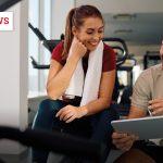 Eine junge Frau mit langem braunen Haar und weißem Handtuch um den Nacken sitzt in Sportkleidung auf einer Bank im Fitnessstudio. Sie lächelt und schaut auf ein Tablet, das ein Mann neben ihr hält. Der Mann trägt ein beiges Poloshirt mit Namensschild und erklärt ihr freundlich etwas auf dem Bildschirm. Beide wirken konzentriert und gut gelaunt. Im Hintergrund sind Fitnessgeräte und große Fenster zu sehen. Oben links steht „SCIENCENEWS“ in weiß-rotem Schriftzug, unten links ist das „fM“-Logo abgebildet.