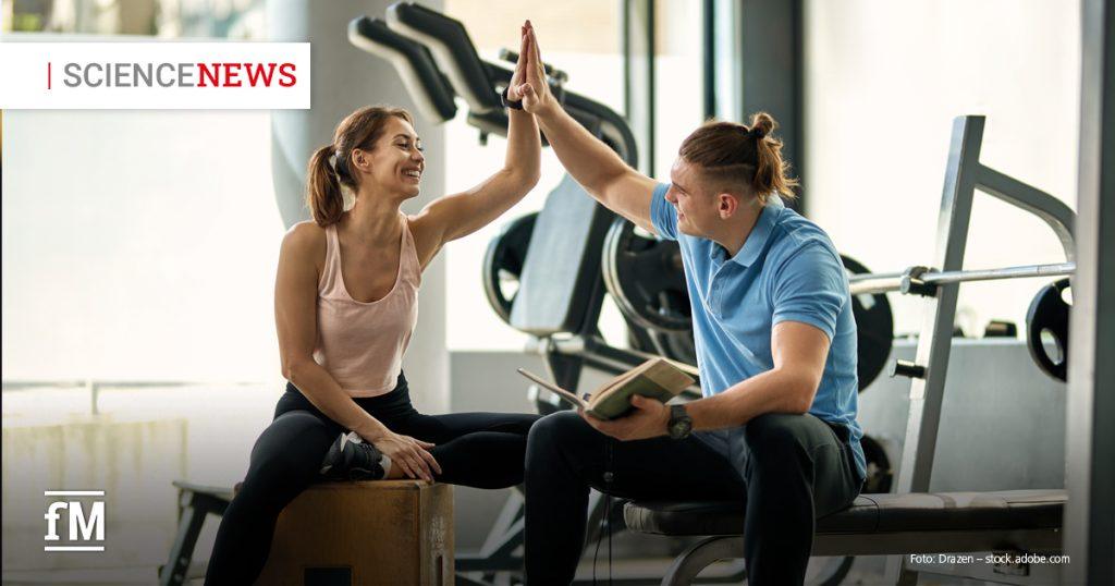 Eine Frau und ein Mann sitzen in einem Fitnessstudio und geben sich lächelnd ein High-Five. Die Frau trägt Sportkleidung und sitzt auf einer Holzkiste, der Mann hält ein Buch in der Hand und sitzt auf einer Hantelbank. Im Hintergrund sind Fitnessgeräte und das Icon 'Science News' zu sehen.