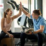 Eine Frau und ein Mann sitzen in einem Fitnessstudio und geben sich lächelnd ein High-Five. Die Frau trägt Sportkleidung und sitzt auf einer Holzkiste, der Mann hält ein Buch in der Hand und sitzt auf einer Hantelbank. Im Hintergrund sind Fitnessgeräte und das Icon 'Science News' zu sehen.