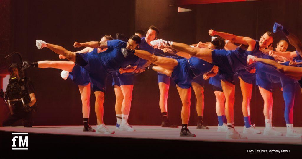 Eine Gruppe von Les Mills Trainern führt auf einer Bühne eine energiegeladene Kampfsport-Choreografie aus. In synchroner Bewegung treten sie mit hoher Präzision und Kraft. Die Teilnehmer tragen blaue Sportkleidung und Boxhandschuhe. Links im Bild filmt ein Kameramann die Performance. Die Szenerie wird durch eine dramatische Bühnenbeleuchtung verstärkt. Das fM-Logo befindet sich unten links.