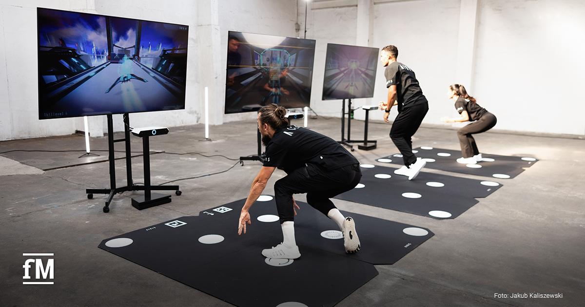 Drei Personen in Sportkleidung machen Übungen auf interaktiven Matten vor Bildschirmen. Die Spiele zeigen futuristische Szenen. Alle tragen schwarze Shirts mit dem Schriftzug 'AIRIS' auf dem Rücken.