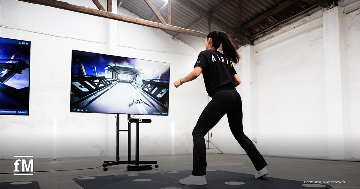 Eine Frau steht in Trainingspose vor einem großen Bildschirm. Darauf ist ein futuristisches Spiel zu sehen. Sie trägt ein schwarzes Shirt mit dem Logo 'AIRIS' und konzentriert sich auf die Bewegung.