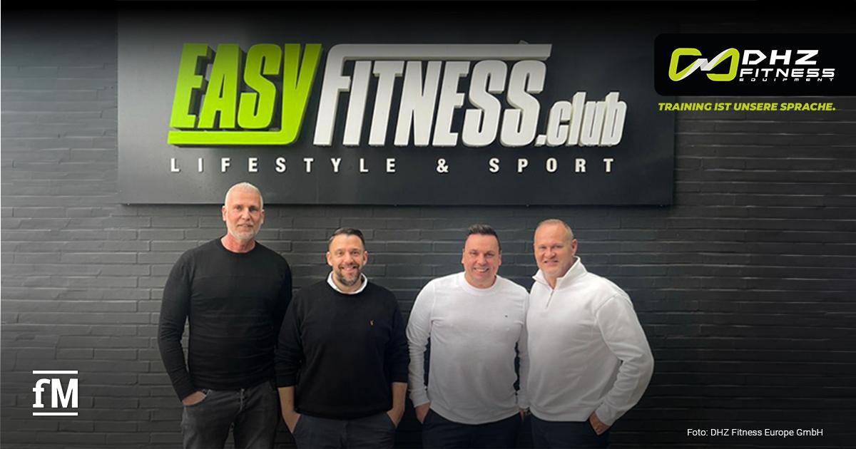 Vier Männer stehen lächelnd vor einer Wand mit dem Logo von EASYFITNESS.club. Sie geben die neue strategische Partnerschaft zwischen DHZ Fitness (das Unternehmenslogo ist in der rechten oberen Bildecke zu sehen) und EASYFITNESS bekannt.