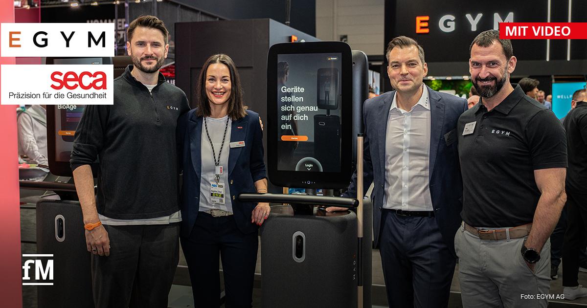 Vier Personen, darunter Mitarbeitende von EGYM und seca, stehen nebeneinander auf der Fitness-Messe FIBO vor einem EGYM-Gerät mit großem Display. Auf dem Bildschirm steht: „Geräte stellen sich genau auf dich ein“. Im Hintergrund sind weitere Messestände und Besucherinnen udn Besucher zu sehen. Links oben befinden sich die Logos von EGYM und seca.