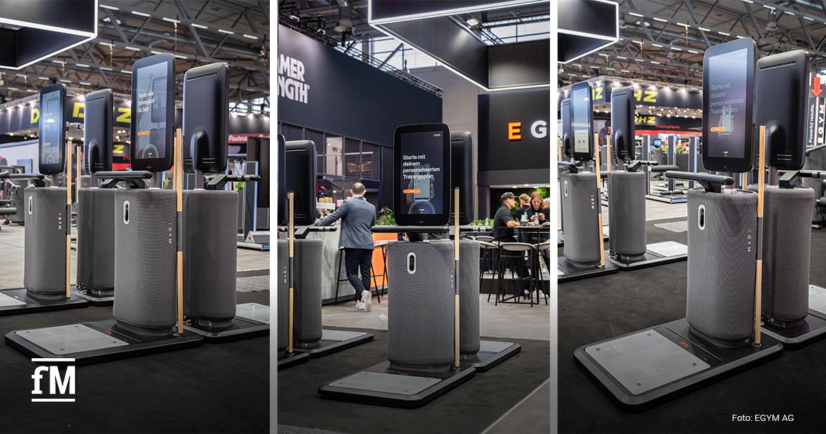 Mehrere EGYM Fitnessgeräte mit großem vertikalem Display stehen nebeneinander auf einer Messefläche. Auf den Bildschirmen sind Aufforderungen wie „Teste wie beweglich du bist“ und „Starte mit deinem personalisierten Trainingsplan“ zu sehen. Besucherinnen und Besucher bewegen sich durch die Messehalle. Die Geräte sind modern gestaltet mit Stoffverkleidung, Holzdetails und einer Bodenplattform.
