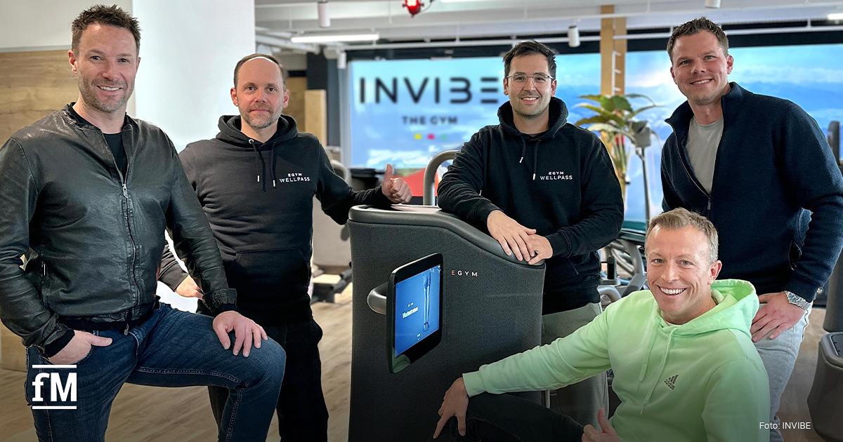 Fünf Männer (Markus Hangl (INVIBE), Hannes Götze (Wellpass), Kim Fischer (Wellpass), Philip Zischke (INVIBE) stehen in einem modernen Fitnessstudio rund um ein Trainingsgerät von EGYM. Florian Streiter (INVIBE) sitzt auf dem Gerät, lächelt und zeigt den Daumen nach oben