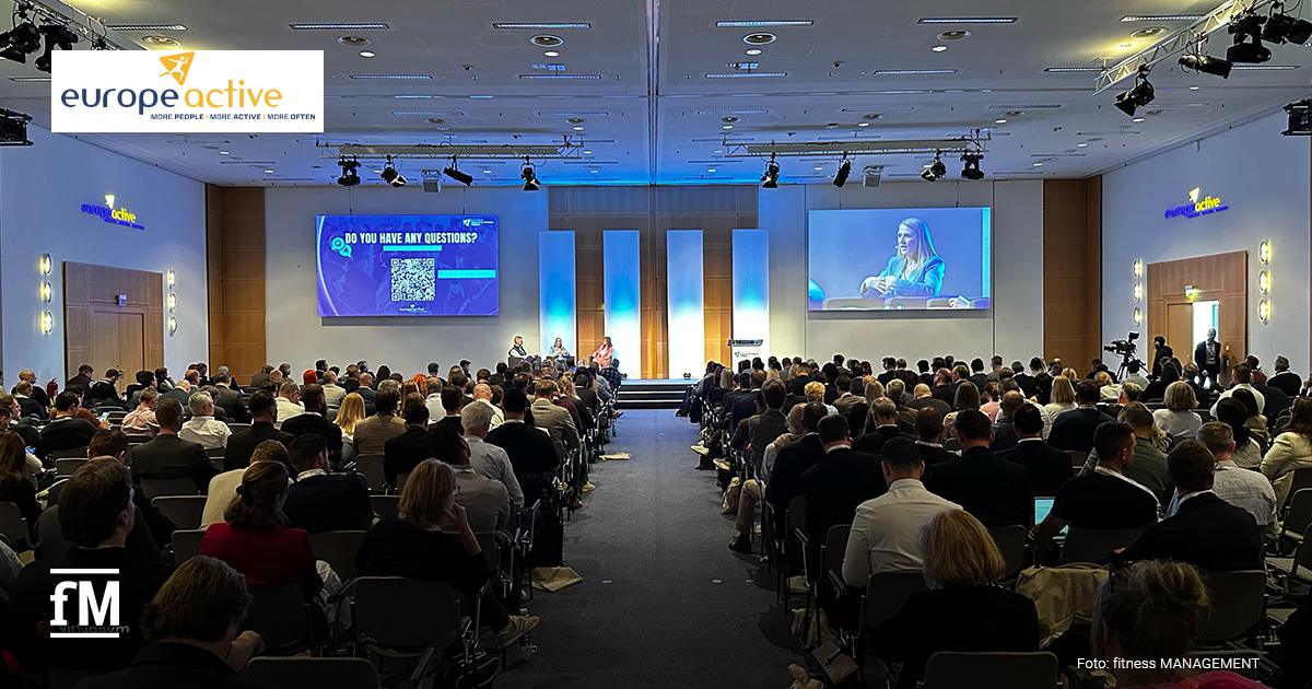 Voll besetzter Konferenzsaal beim European Health & Fitness Forum 2025 in Köln – Bühne mit Podiumsdiskussion, zwei großen Screens, darunter eine Frageaufforderung und eine Rednerin im Großbild. Logos von EuropeActive und fitness MANAGEMENT sichtbar.
