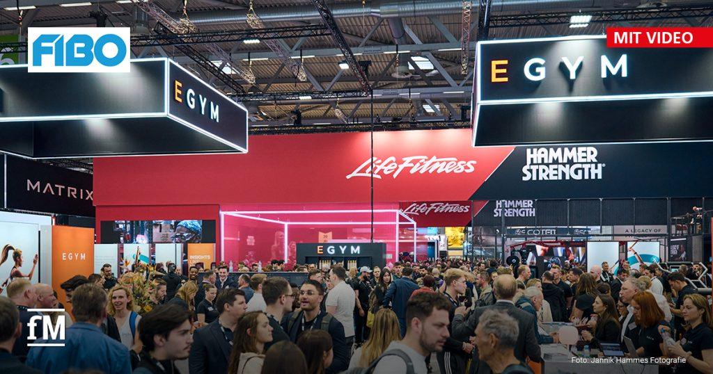 Dichtes Gedränge auf der Messe FIBO. Zu sehen sind große Stände von 'EGYM', 'Life Fitness' und 'Hammer Strength'. Besucher tauschen sich aus, schauen Geräte an oder machen Fotos. Rechts oben steht 'Mit Video'.