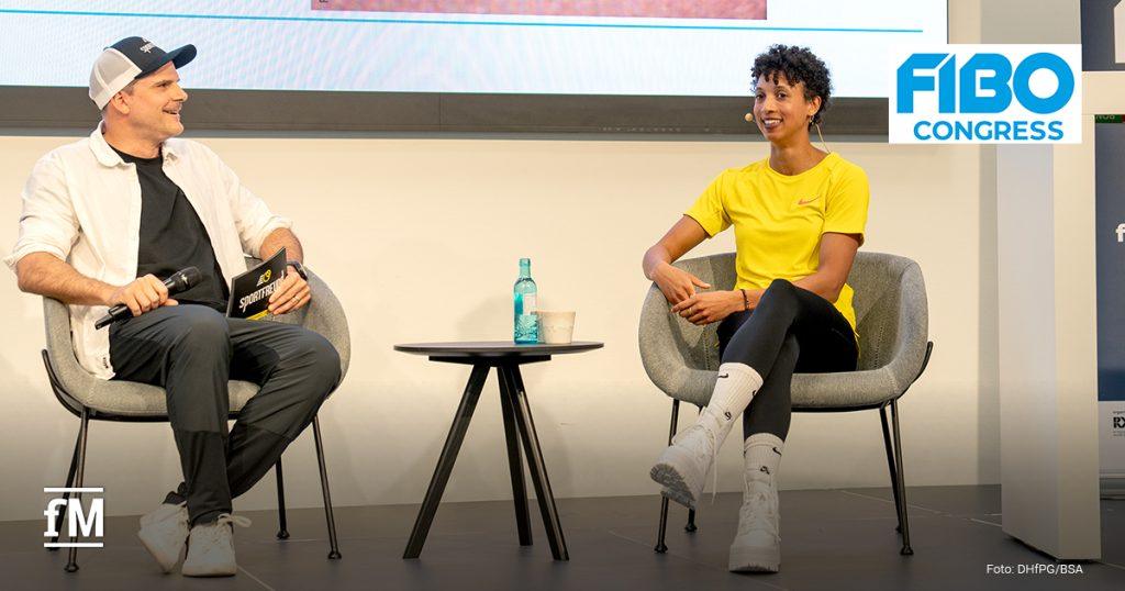 Eine Frau (Olympiasiegerin Malaika Mihambo, rechts) und ein Mann (Triathlonweltmeister Daniel Unger, links) sitzen auf Sesseln und führen ein Bühneninterview beim 'FIBO Congress', im Hintergrund ist der Anschnitt einer Powerpointpräsentation zu sehen. Die Frau trägt ein gelbes Shirt, der Moderator hält Mikrofon und Karte.