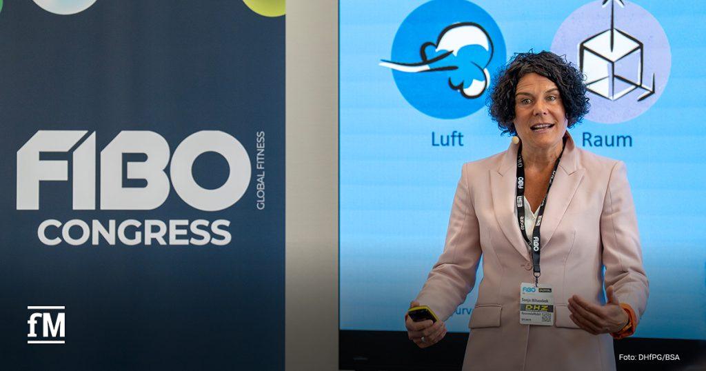 Eine Frau im rosa Blazer spricht beim 'FIBO Congress'. Hinter ihr sind Symbole für 'Luft' und 'Raum' zu sehen. Sie trägt ein Namensschild und ein Headset-Mikrofon.