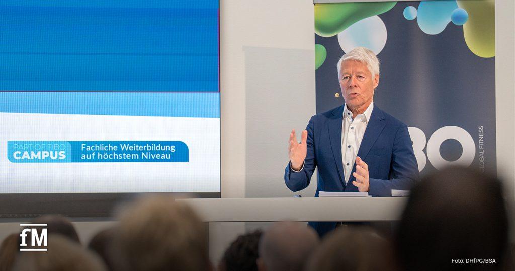 Ein älterer Mann im blauen Anzug spricht auf einer Bühne beim 'FIBO Congress'. Neben ihm steht: 'Fachliche Weiterbildung auf höchstem Niveau'. Er spricht engagiert ins Publikum.