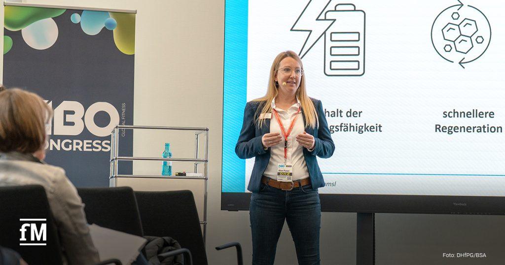Eine Frau hält beim 'FIBO Congress' einen Vortrag. Sie trägt Blazer, Brille und Mikrofon. Hinter ihr ist eine Präsentation mit Icons zu Energie, Leistungsfähigkeit und Regeneration zu sehen.