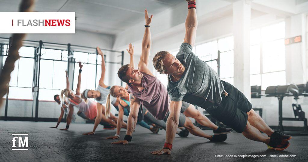 Auf dem Bild sind mehrere Frauen und Männer zu sehen, die in einem Fitnessstudio seitlich gestützte Plank-Übungen ausführen. Sie heben dabei jeweils einen Arm nach oben und haben Spaß beim Workout. Im Hintergrund sind große Fenster und Trainingsgeräte zu sehen.