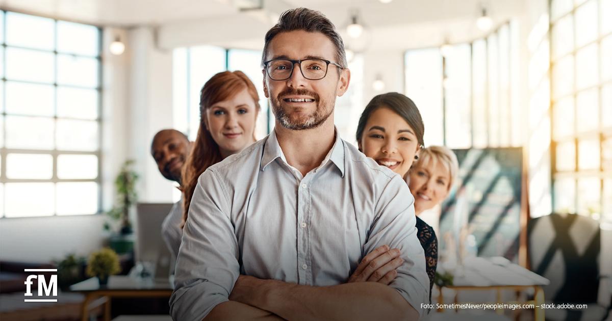 Fünf lächelnde Personen unterschiedlicher Herkunft stehen eng beieinander in einem hellen Büro, vorne ein Mann mit Brille und verschränkten Armen.