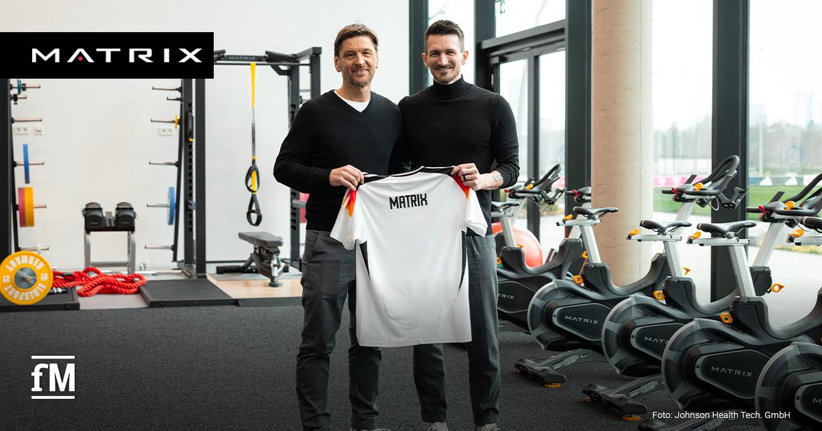 DFB-Athletiktrainer Nicklas Dietrich (rechts) und Volker Lichte (links) stehen im modernen Fitnessraum des DFB-Campus und halten gemeinsam ein weißes Deutschland-Trikot, oben auf der Rückseite der schwarze Aufdruck in Großbuchstaben 'Matrix'. Im Hintergrund sind Trainingsgeräte von Matrix Fitness zu sehen.