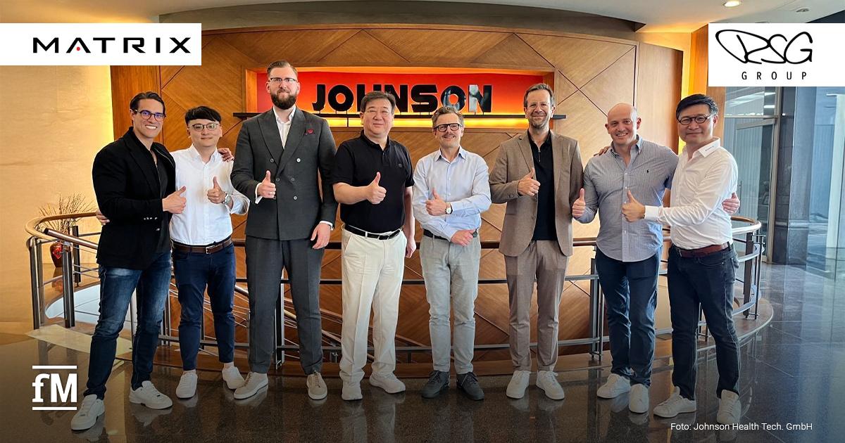 Auf dem Bild ist eine Delegation von Johnson Health Tech. und der RSG Group zu sehen. Acht Männer stehen nebeneinander vor einem Holz-Hintergrund mit dem Schriftzug „JOHNSON“. Sie lächeln und zeigen Daumen hoch. Alle tragen Business- oder Business-Casual-Outfits. Im Bildhintergrund ist eine geschwungene Treppe zu sehen.