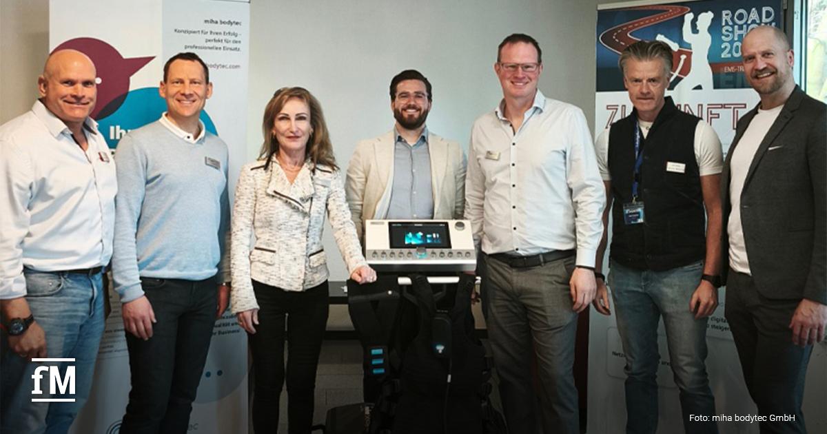 Teilnehmende der EMS-Roadshow in Düsseldorf stehen nebeneinander in einem Raum und lächeln in die Kamera. In der Mitte befindet sich ein EMS-Gerät (Elektromuskelstimulation) mit Weste. Hinter der Gruppe sind zwei Roll-up-Banner zu sehen – einer davon mit dem Logo von „miha bodytec“, der andere mit der Aufschrift „Roadshow 2024“. Die Gruppe besteht aus Männern und einer Frau, alle in Business- oder Smart-Casual-Kleidung.