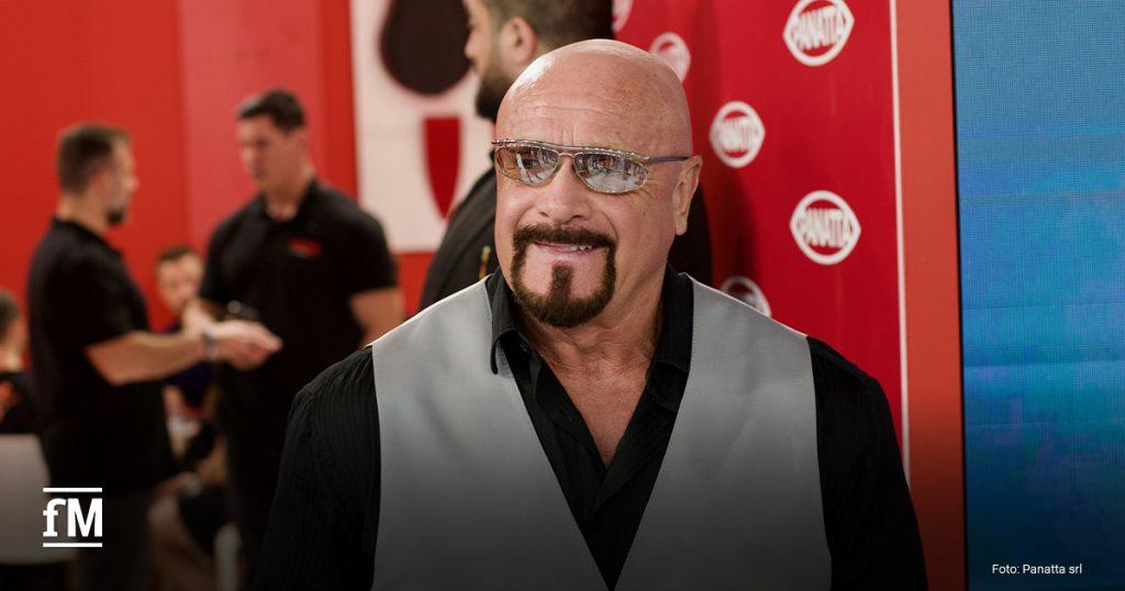 Rudy Panatta steht vor dem roten FIBO-Messestand. Er trägt ein schwarzes Hemd, eine grauen Anzugweste und eine Brille mit verspiegelten Gläsern. Er lächelt leicht und blickt aus dem Bild heraus.