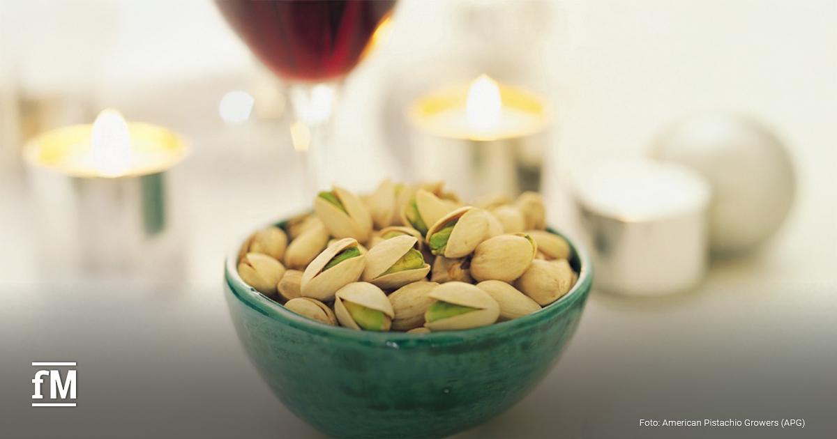 Stimmungsvolle Szene mit einer grünen Schale voller Pistazien, daneben ein Glas Rotwein, Kerze und Serviette auf einem gedeckten Tisch.