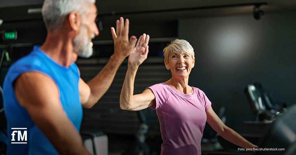 Ein älteres Paar trainiert gemeinsam im Fitnessstudio. Beide laufen auf Laufbändern und geben sich lächelnd ein motivierendes High-Five. Die Stimmung ist fröhlich und aktiv, der Raum ist dezent beleuchtet und mit Trainingsgeräten ausgestattet