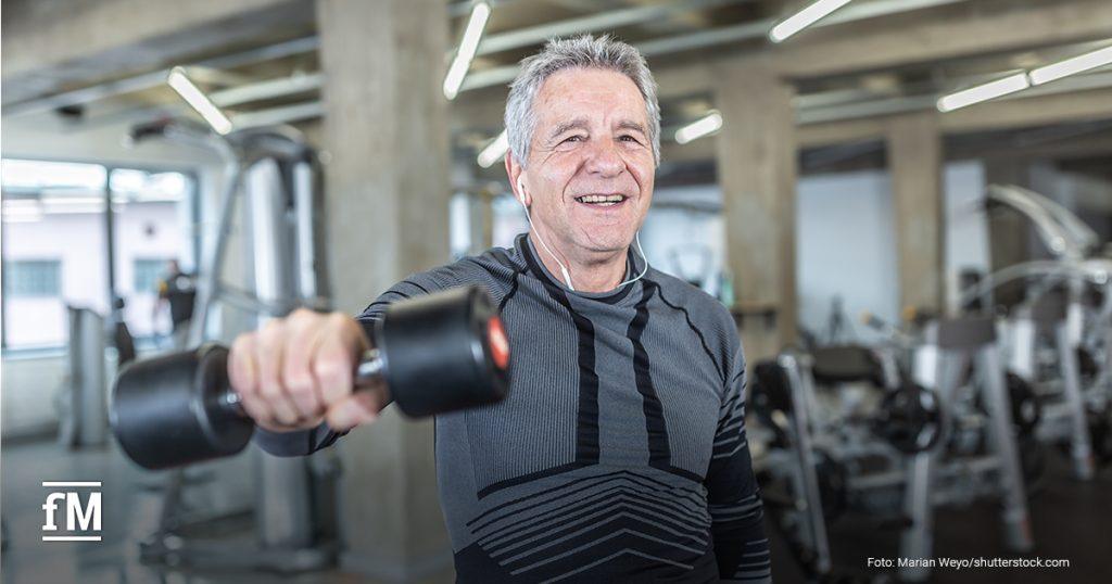 Ein älterer Mann trainiert mit einer Kurzhantel im Fitnessstudio. Er trägt ein sportliches Langarmshirt, lächelt motiviert in die Kamera und hört über Kabelkopfhörer Musik. Im Hintergrund sind verschiedene Trainingsgeräte zu sehen