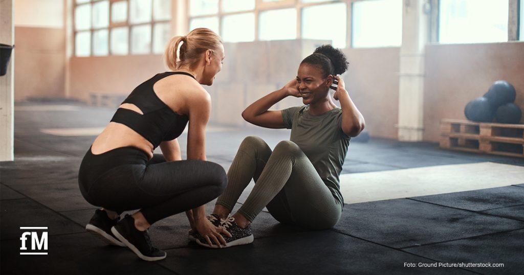 wei junge Frauen trainieren gemeinsam in einem Fitnessstudio. Eine Frau führt Sit-ups aus, während die andere ihre Füße fixiert und sie motivierend unterstützt. Beide lächeln und wirken konzentriert. Im Hintergrund sind Gymnastikbälle und Paletten zu sehen