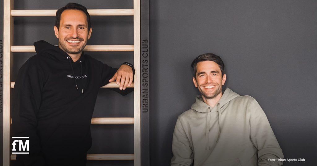 Zwei Männer in sportlichen Kapuzenpullovern lächeln in die Kamera. Moritz Kreppel (CEO und Co-Founder von Urban Sports Club) lehnt an einer Sprossenwand mit dem Logo „Urban Sports Club“, Benjamin Roth sitzt entspannt daneben.