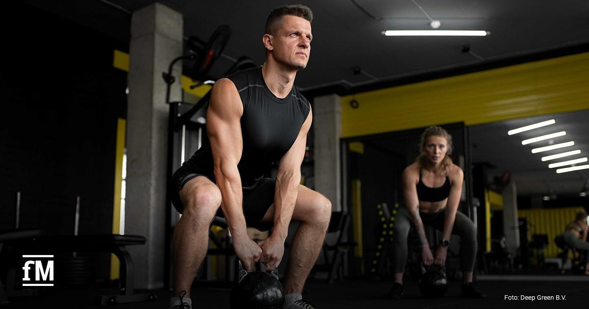 Zwei sportlich trainierte Personen beim intensiven Kettlebell-Workout in einem modernen Fitnessstudio. Links im Vordergrund steht ein Mann mittleren Alters, rechts eine junge Frau