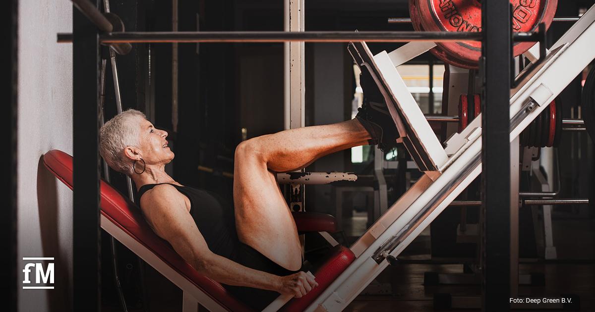 Eine ältere, sportlich wirkende Frau mit kurzen grauen Haaren trainiert in einem Fitnessstudio an einer Beinpresse. Sie trägt einen schwarzen Trainingsanzug und wirkt konzentriert und motiviert.