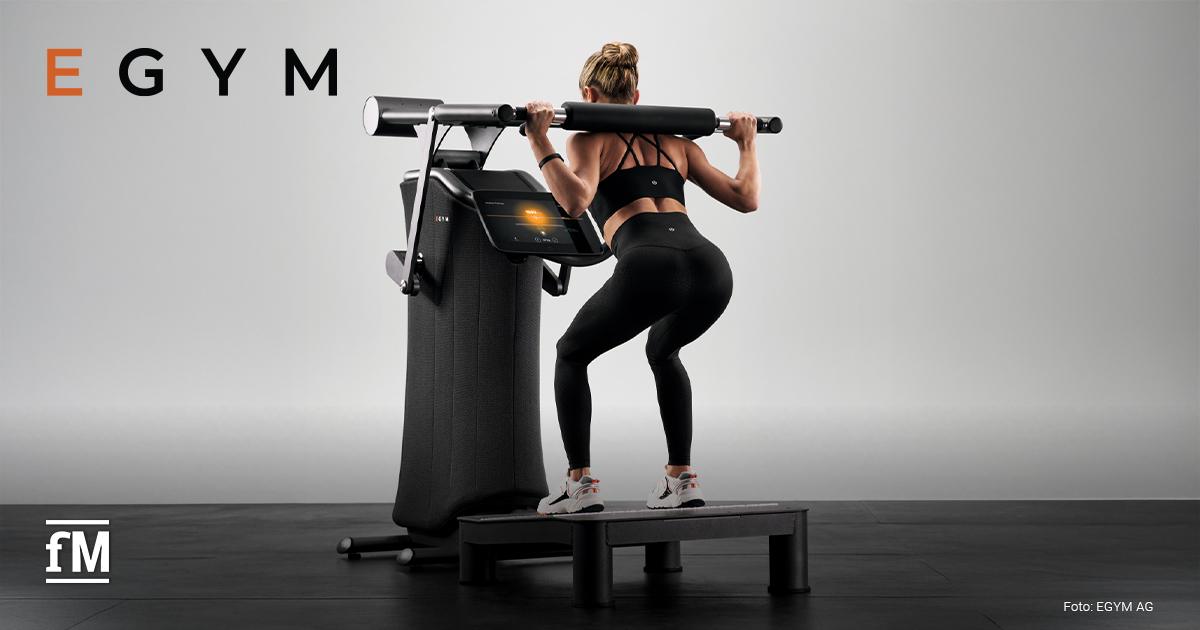 Eine Frau in Sportkleidung macht Kniebeugen an einem modernen EGYM-Trainingsgerät mit Display in einem hellen Fitnessraum.