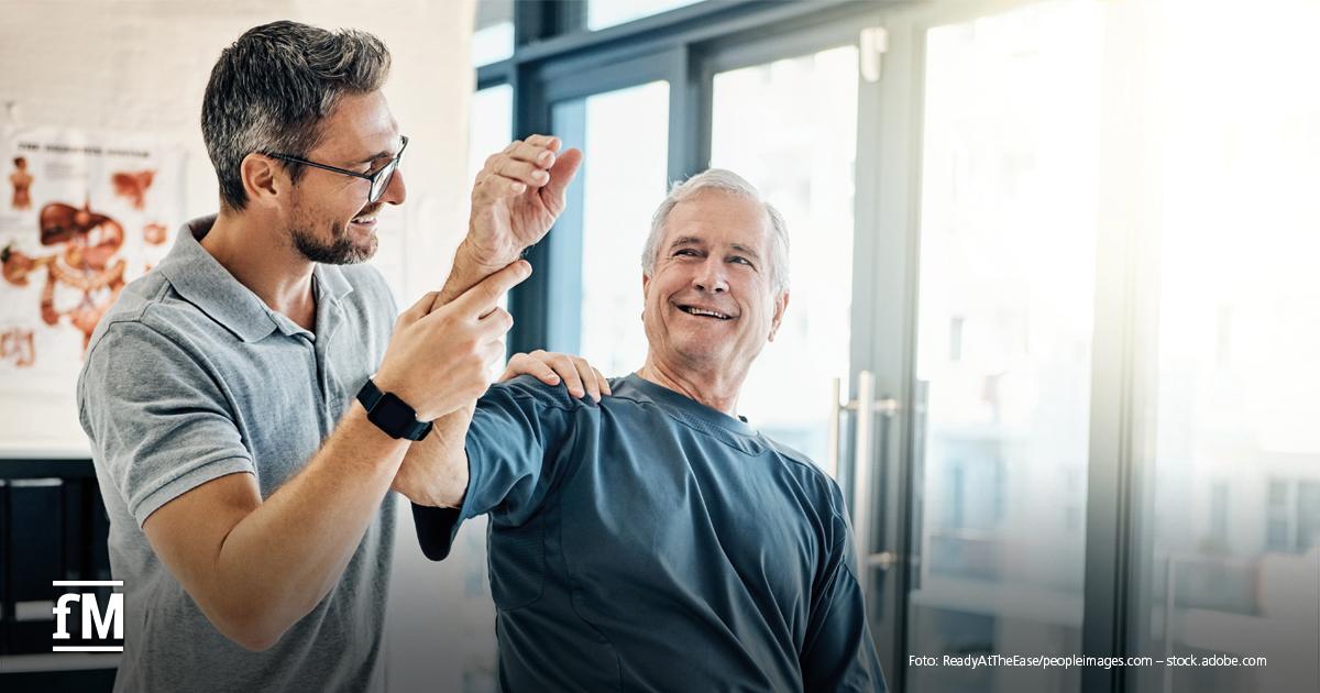Ein älterer Mann in Sportkleidung sitzt in einem hellen Behandlungsraum. Ein jüngerer Mann unterstützt ihn freundlich dabei, seinen rechten Arm anzuheben – vermutlich im Rahmen einer physiotherapeutischen Übung. Beide lachen, im Hintergrund ist eine Fensterfront zu sehen. Links unten befindet sich das fM-Logo.